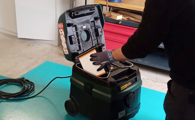 Eine Person mit Handschuhen öffnet den Deckel eines schwarzen Staubsaugers. Der Staubsauger steht auf einem blauen Untergrund. Die Person hält einen weißen Filter, der gerade eingesetzt wird. Im Hintergrund sind holzfarbene Kisten und eine Wand zu sehen.